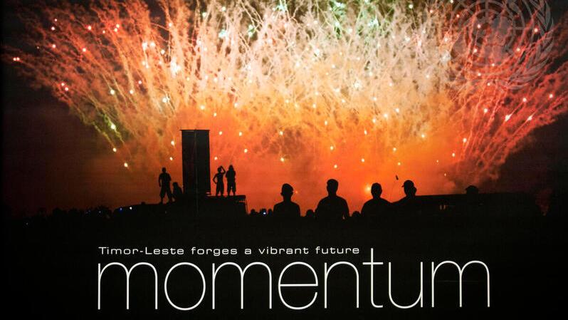 Silhouetted figures watch colorful fireworks light up the night sky in Timor-Leste, with the words "Timor-Leste forges a vibrant future – momentum" overlaid on the image.