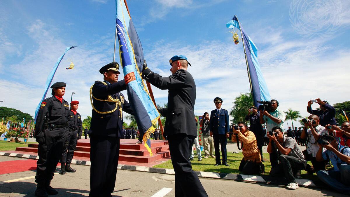 A UN peacekeeper hands over a ceremonial flag to a Timor-Leste police officer during a formal event, as photographers and onlookers capture the moment under a clear blue sky.