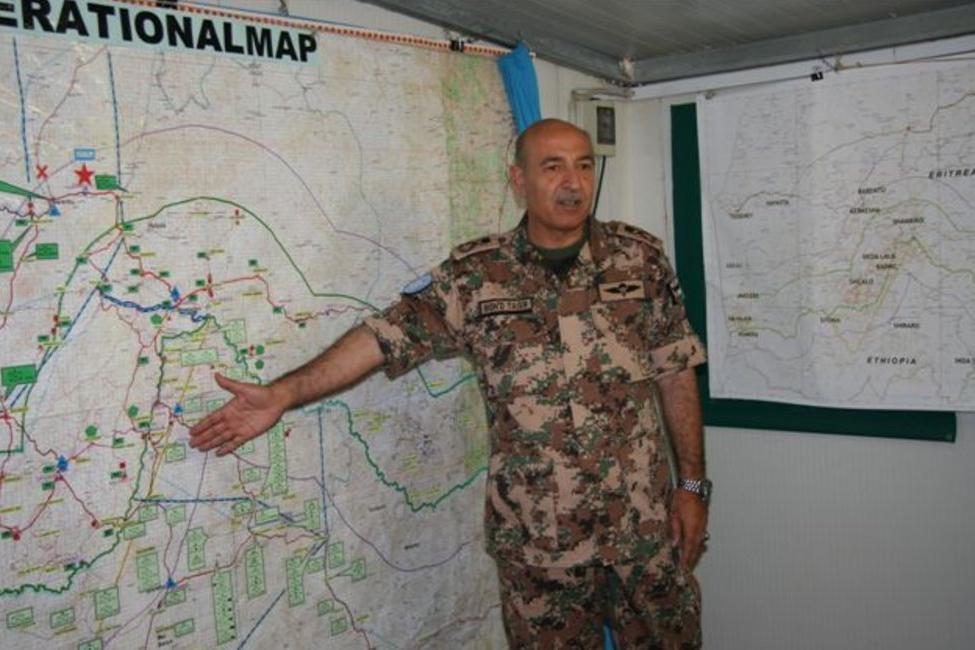 A man in military uniform standing in front of a large map indicating a section of the map with an open hand.