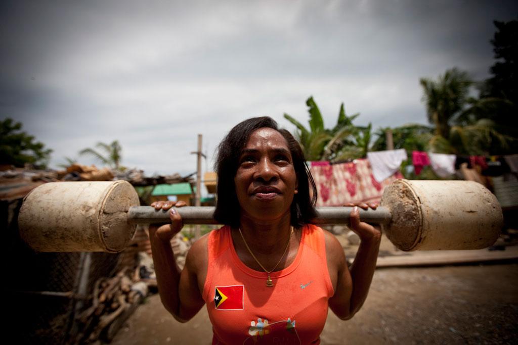 An athlete lifts a heavy barbell across the shoulders outdoors, standing in a residential area with trees, buildings, and laundry visible in the background.
