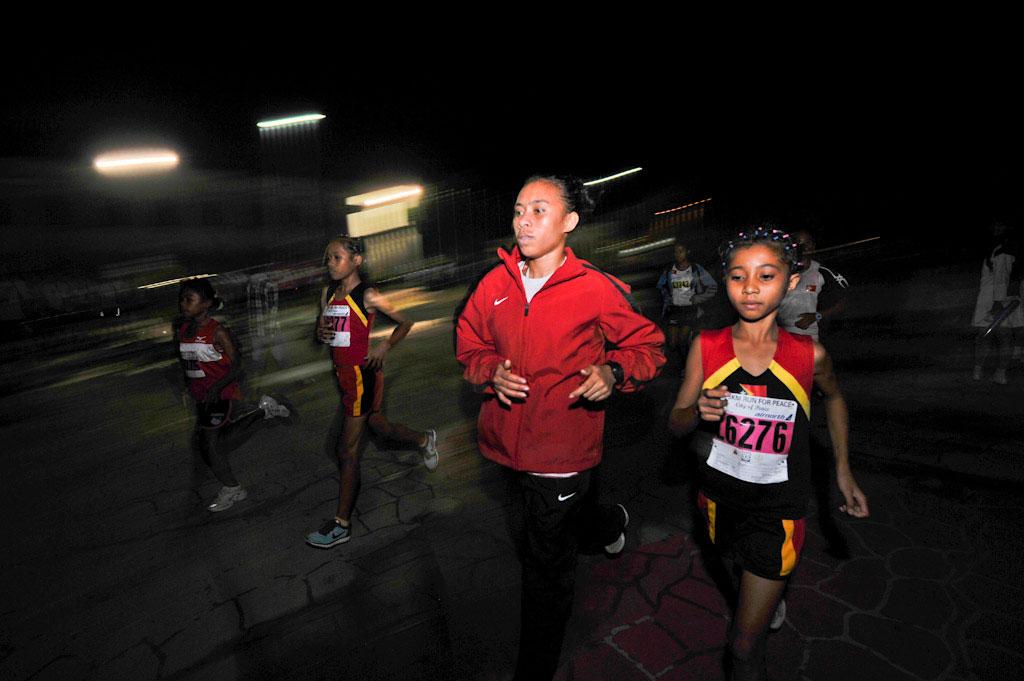 Runners participate in a night road race, moving along a dim<lluminated street. Several participants wear numbered bibs as they run together in a group.