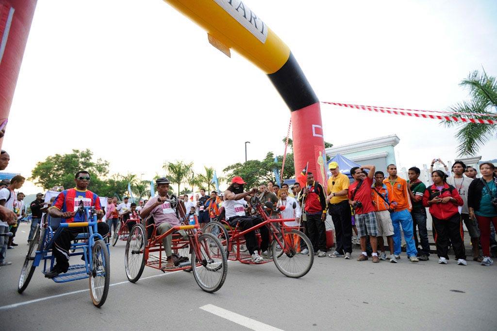 A road race featuring athletes using hand‑powered tricycles at a starting line, with spectators watching from both sides of the street beneath an inflatable race arch.