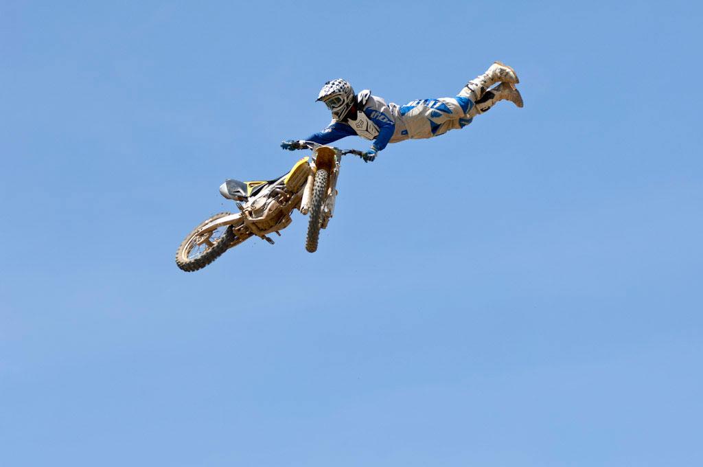 A motocross rider performs an aerial stunt, stretched horizontally while holding a dirt bike high above the ground against a clear blue sky.