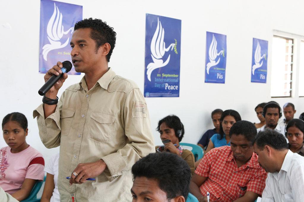A man speaking into a microphone surrounded by an audience. In the background are posters promoting International Day of Peace.