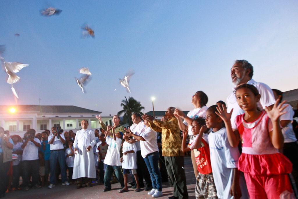 A group of people congregate as doves fly into the sky.